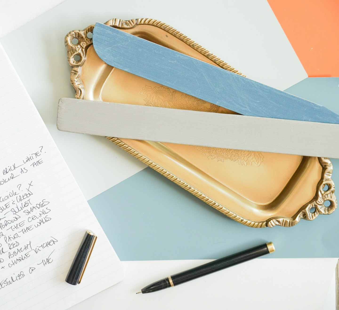 Notebook and pen next to painted wood samples on a desk.